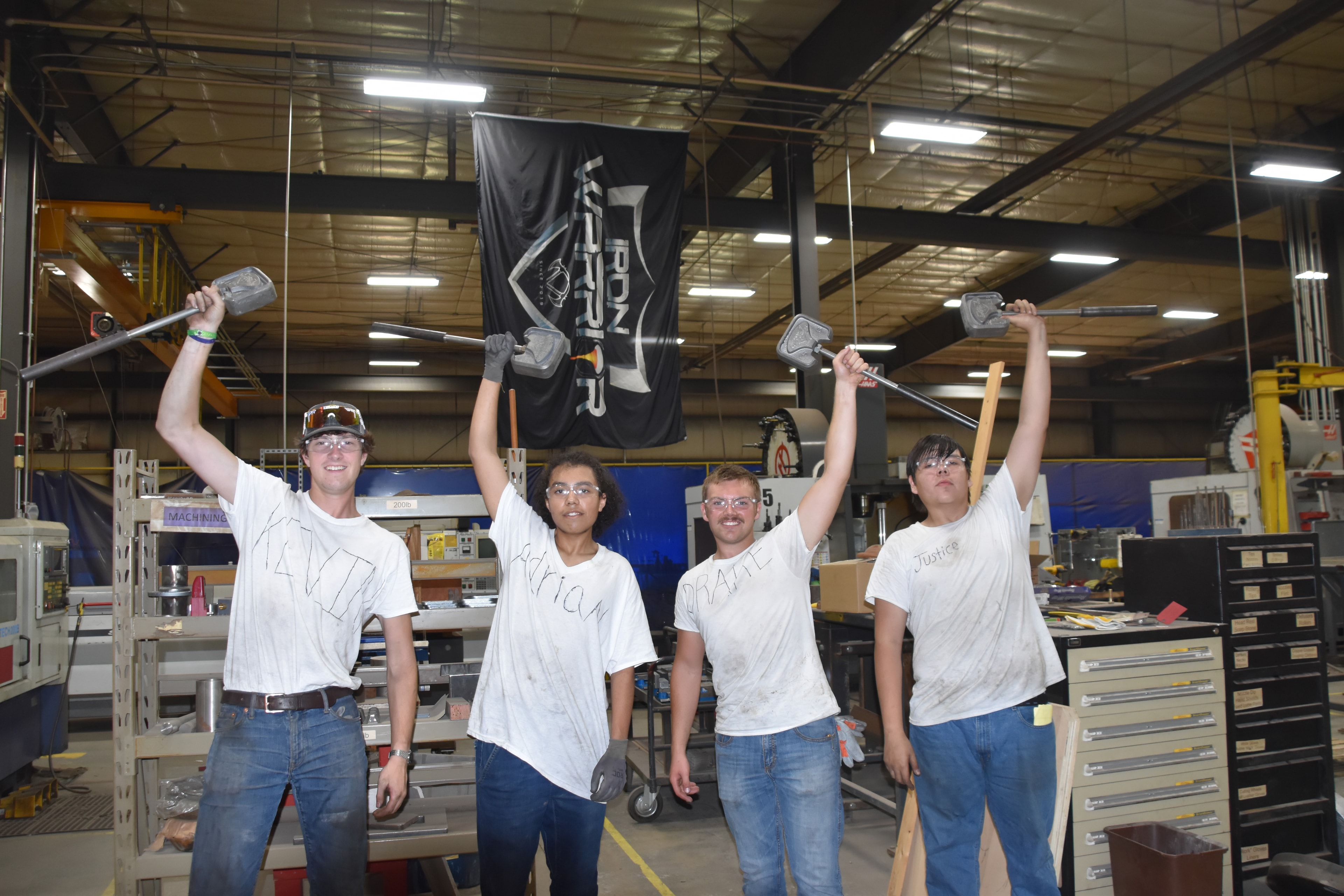 Four Iron Warrior Academy students in a foundry workshop, raising ladles in celebration after completing a casting exercise
